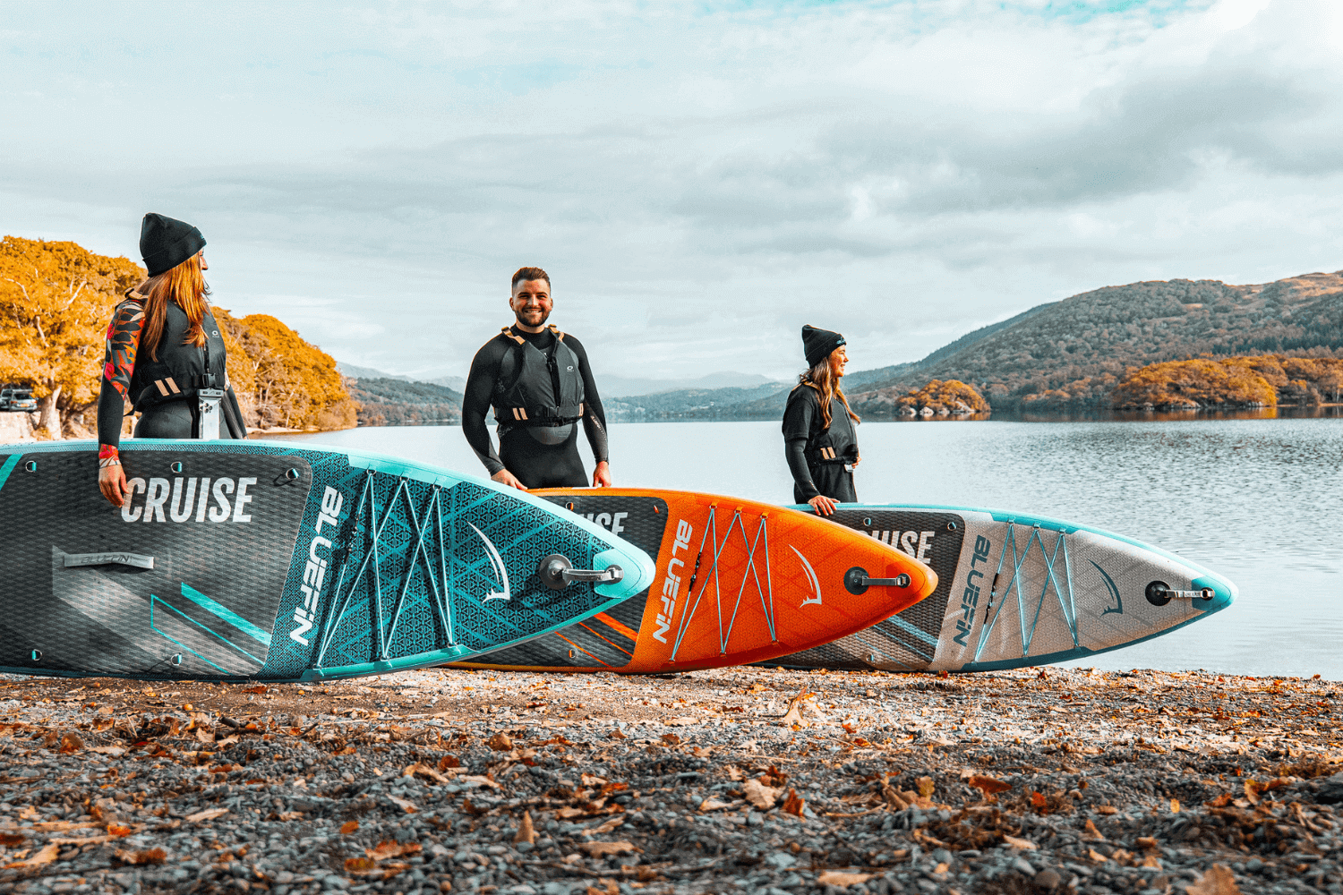 Three people standing by the water with inflatable paddleboards in blue, orange, and gray colors.