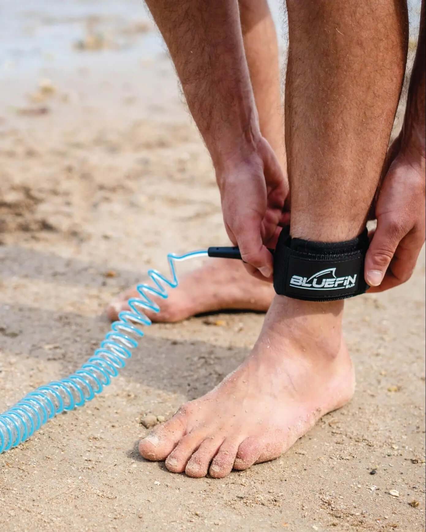 Person securing a black ankle strap with a blue coiled leash on sandy beach