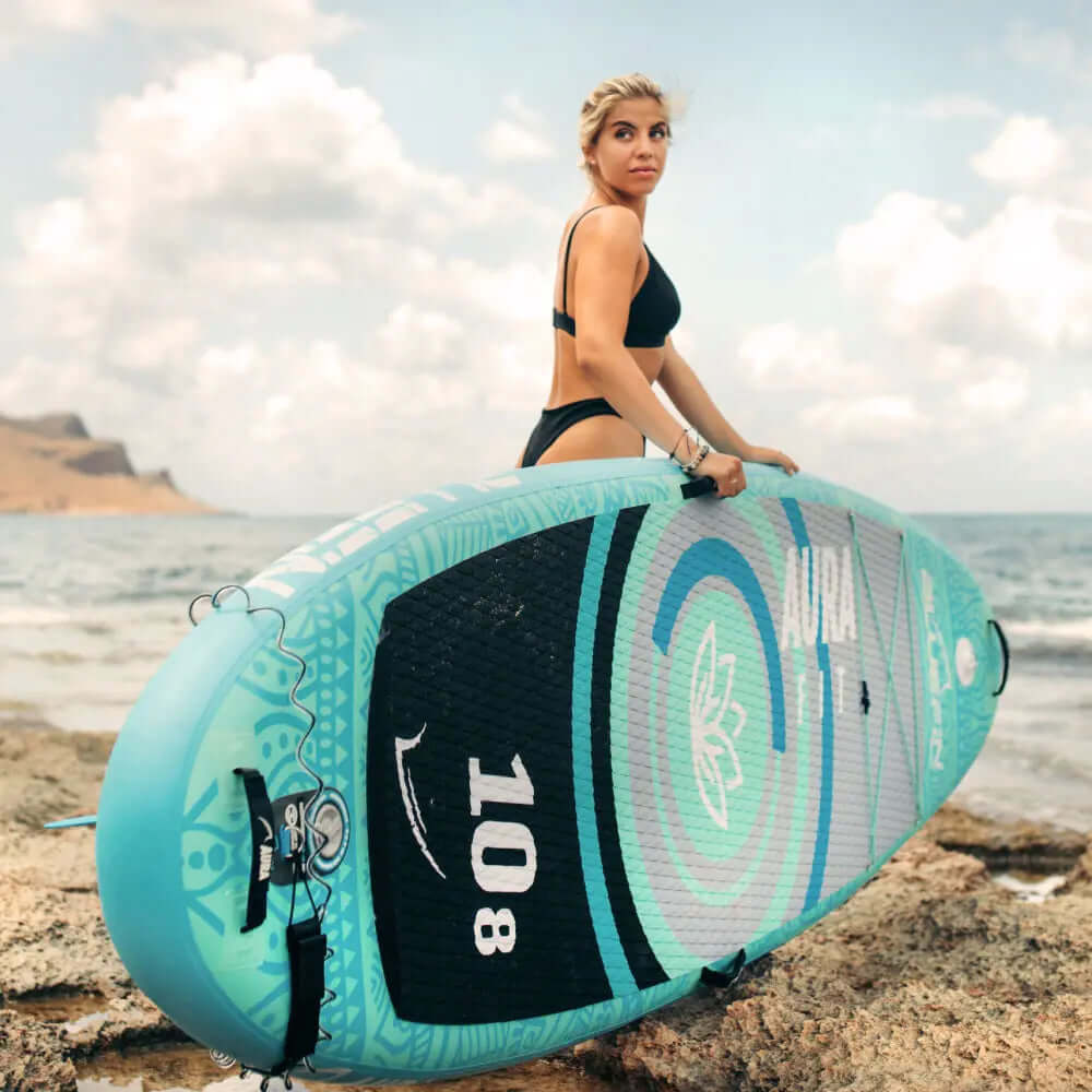 Woman in a black bikini holding a turquoise inflatable paddleboard by the shore with ocean waves.
