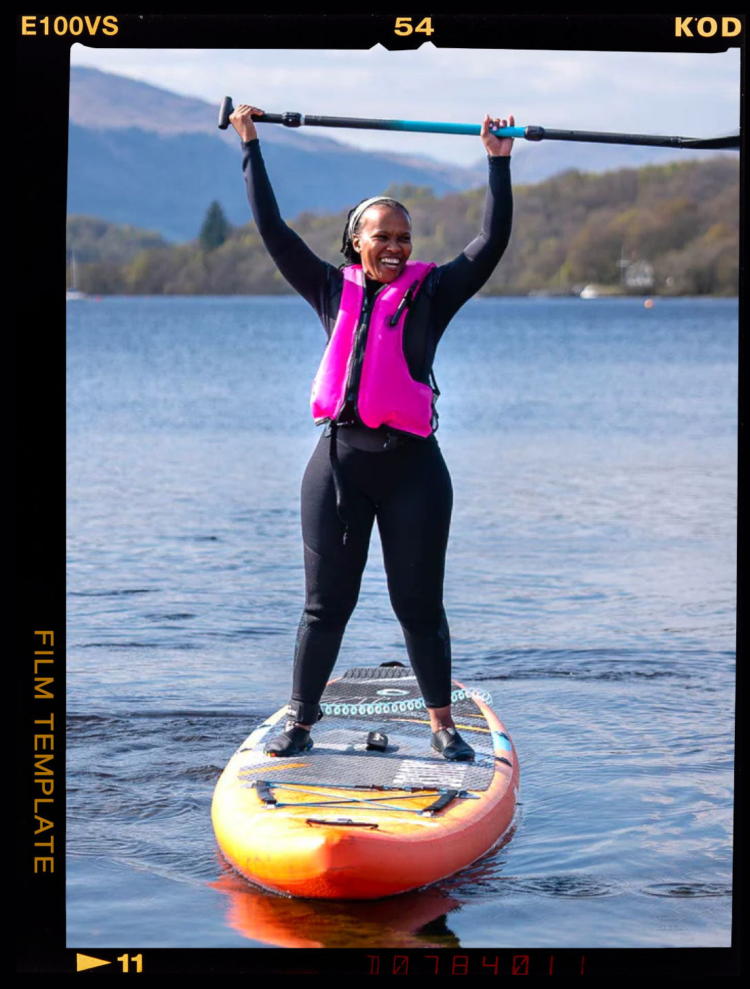 Woman in a pink life vest stands on an orange paddleboard, holding a paddle above her head, smiling.