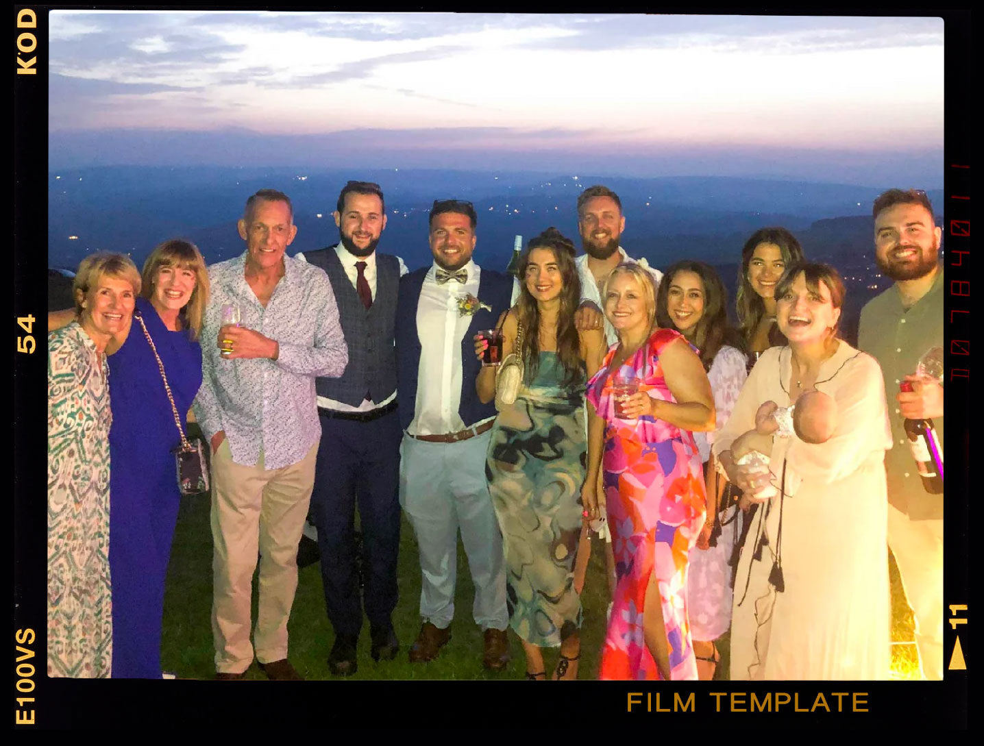 Group of 12 people smiling outdoors at sunset, wearing formal and semi-formal attire, holding drinks.