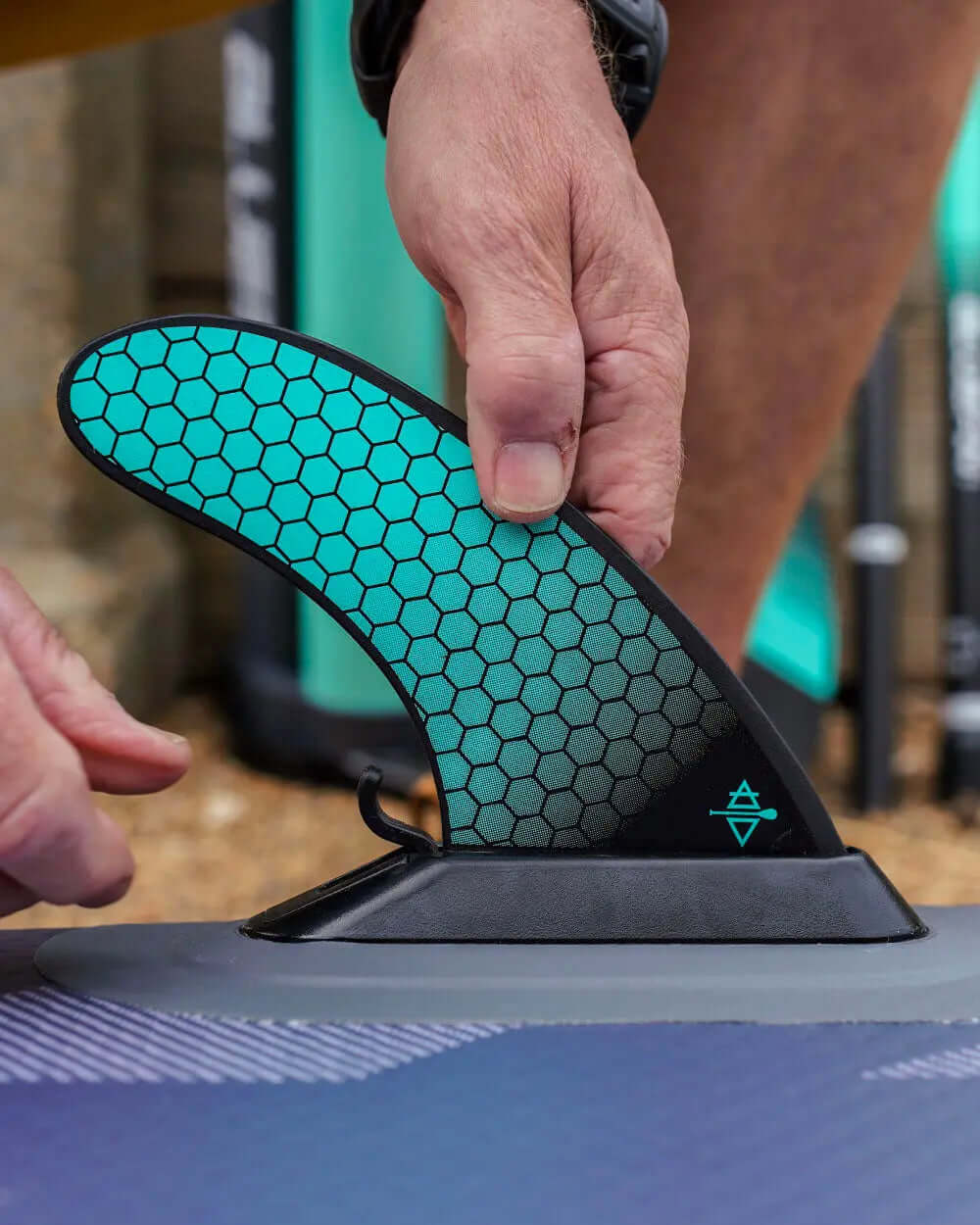 Person attaching a black and teal hexagonal fin to a paddleboard on a sandy surface