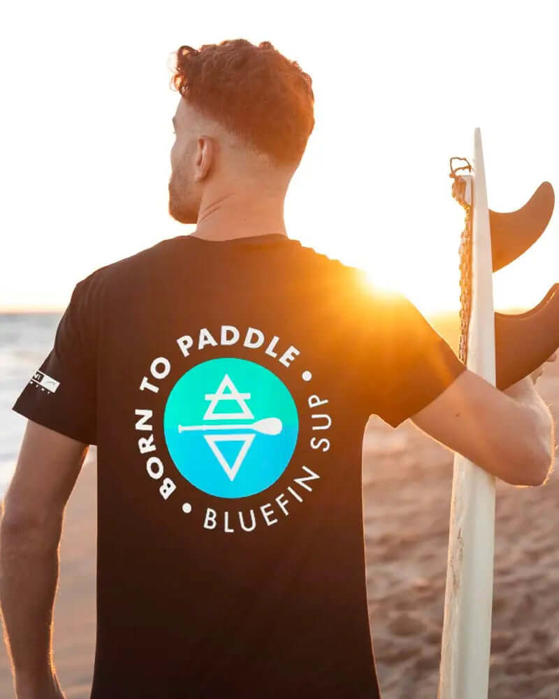 Person wearing a black t-shirt with 'Born to Paddle' and Bluefin SUP logo, holding a paddleboard.