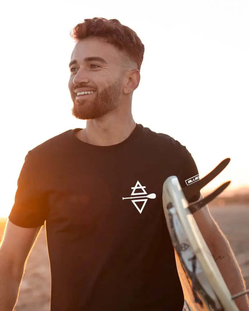 Man smiling in a black t-shirt with a logo, holding a surfboard at sunset on the beach