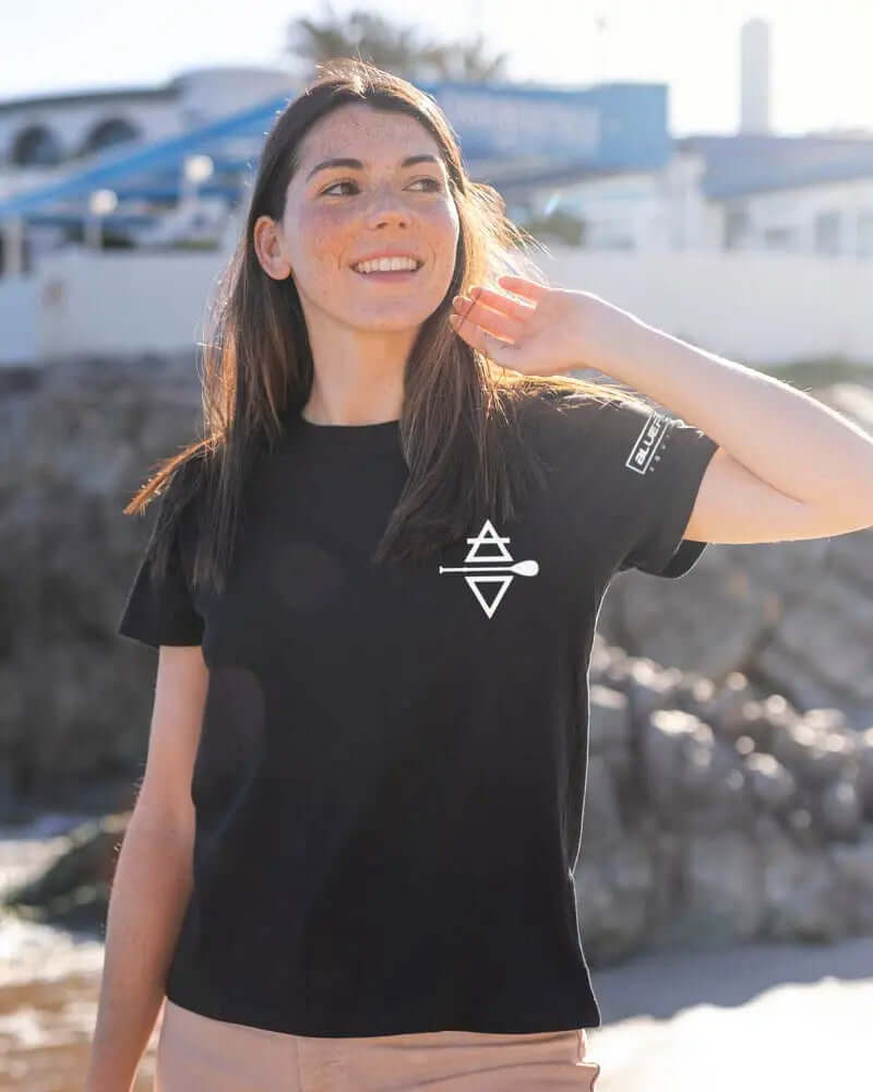 Woman in a black t-shirt with a white logo, smiling and touching her hair by the beach.
