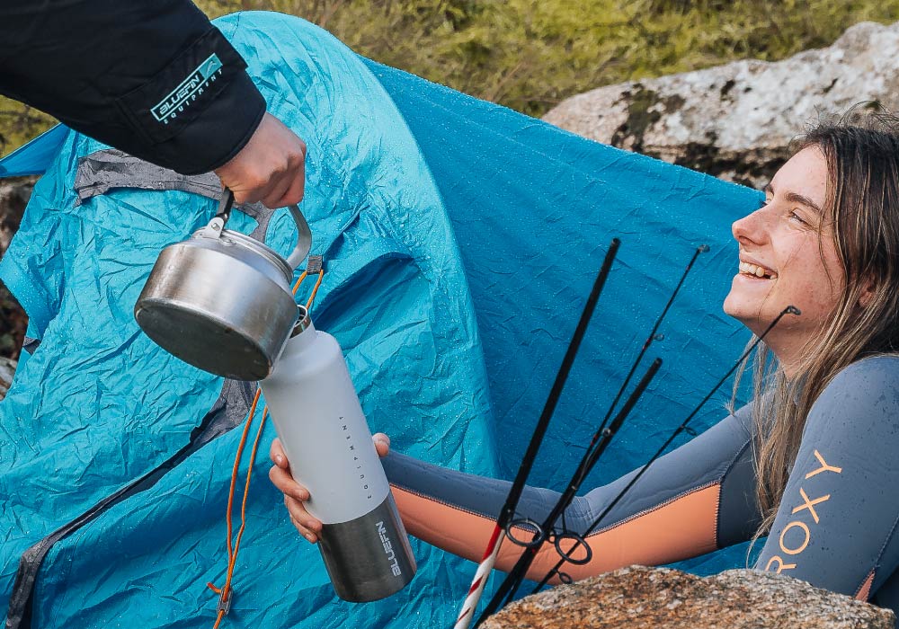 Person in wetsuit smiling while receiving a kettle from another person near a blue tent outdoors.