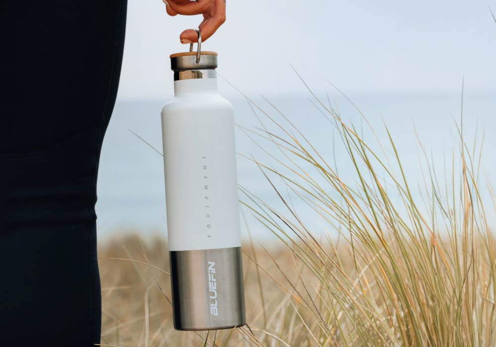Person holding a white and stainless steel water bottle with a wooden lid in a grassy area near the beach.