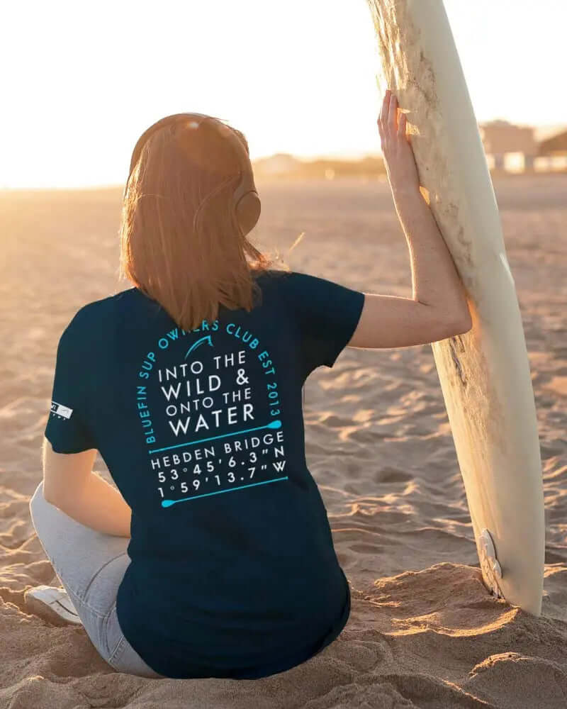 Person sitting on sand wearing navy t-shirt with text and graphics, holding a surfboard.