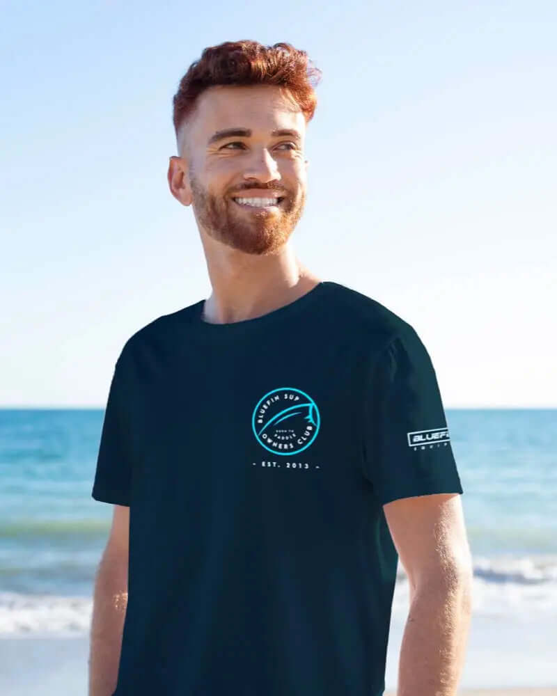 Man smiling on beach wearing navy Bluefin SUP t-shirt with logo on front and sleeve