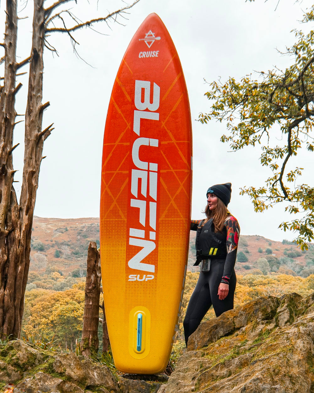 Inflatable Bluefin SUP paddleboard in orange and yellow, woman in wetsuit standing beside it outdoors.