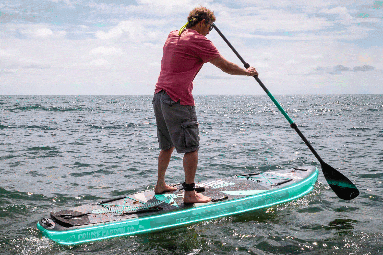Person paddling on a turquoise inflatable paddleboard in the ocean under a cloudy sky