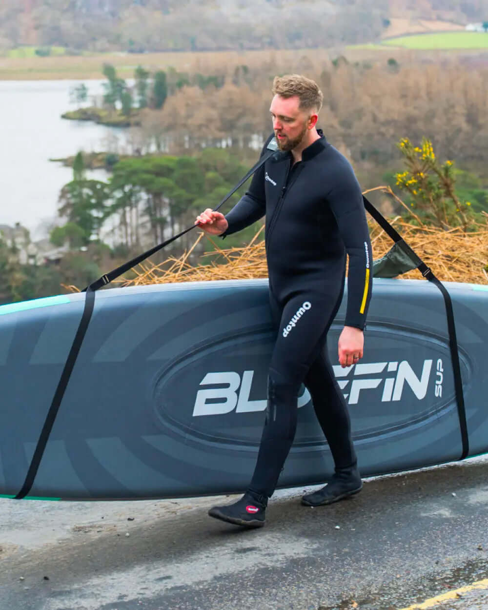 Man in a wetsuit carrying a Bluefin SUP paddleboard along a path by a lake.