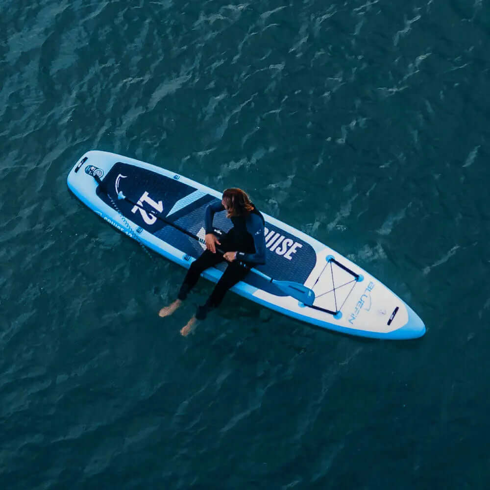 Person sitting on a blue inflatable paddleboard in calm water, wearing a wetsuit.