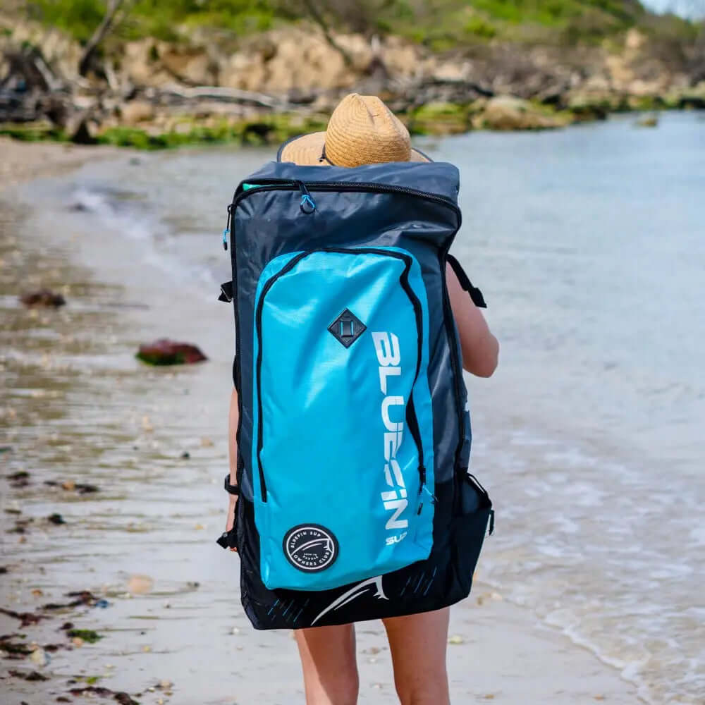 Person walking on the beach carrying a large blue and black Bluefin SUP backpack.