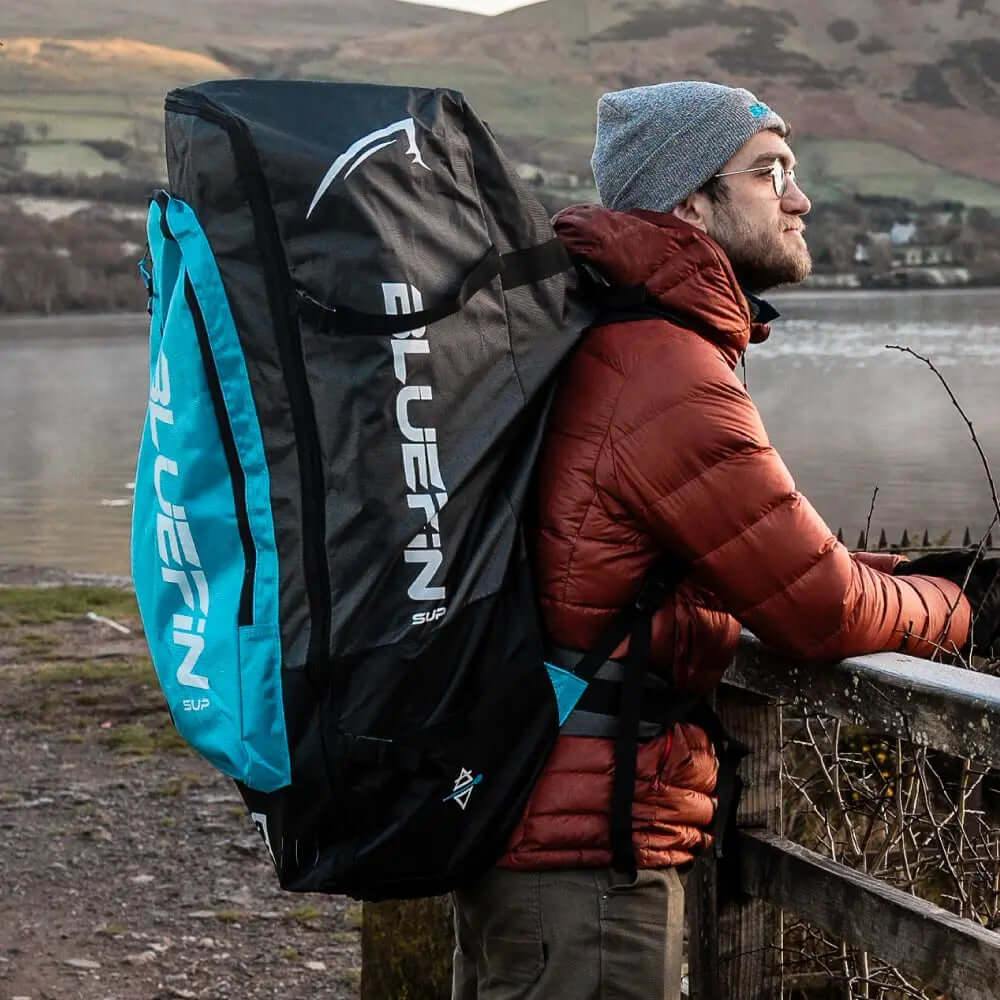 Man wearing a red jacket and gray beanie, carrying a Bluefin SUP backpack by a lake.