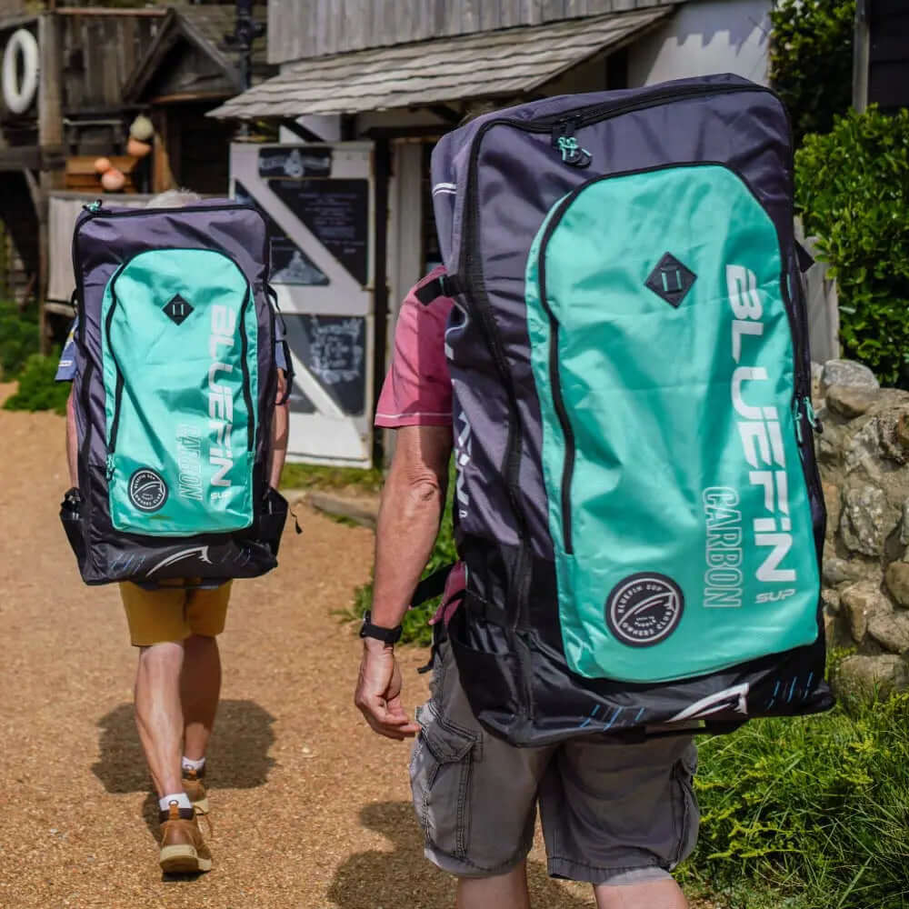 Zwei Personen gehen mit großen Bluefin Carbon SUP Rucksäcken auf einem Kiesweg in der Nähe von Gebäuden.