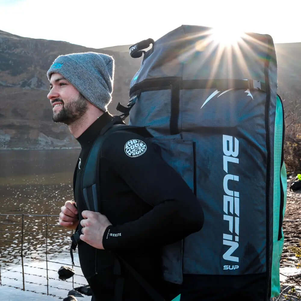 Mann im Neoprenanzug, der einen großen Bluefin SUP Rucksack an einem See mit Bergen im Hintergrund trägt.
