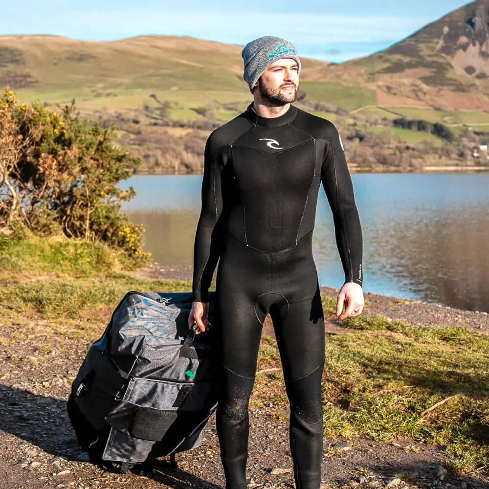 Mann im schwarzen Neoprenanzug, der eine Rolltasche an einem See mit Hügeln im Hintergrund trägt.