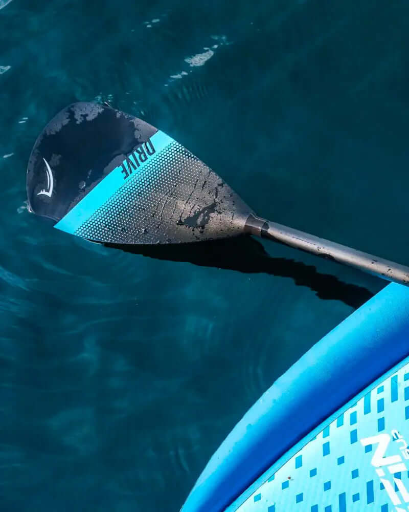 Paddle with black and blue design resting on a blue inflatable paddleboard in clear water