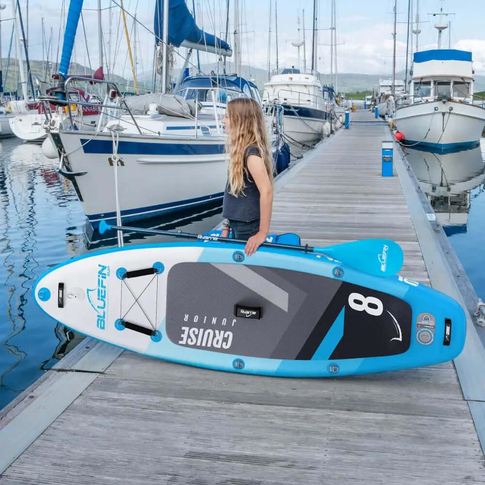 Child holding a Bluefin inflatable paddleboard on a dock near boats, with calm water reflections.