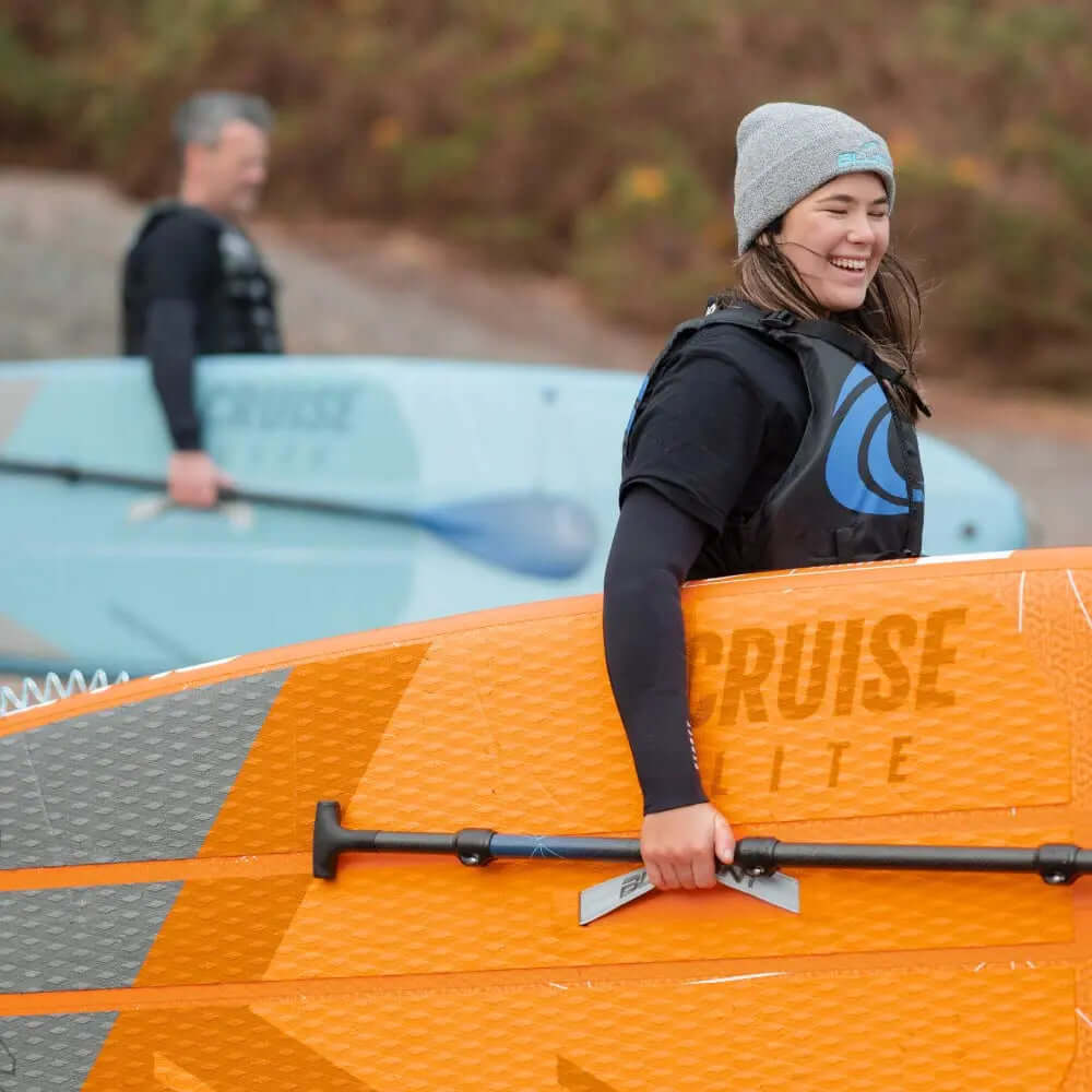 Two people at the beach with an orange paddleboard and a light blue paddleboard, smiling and preparing to paddle.