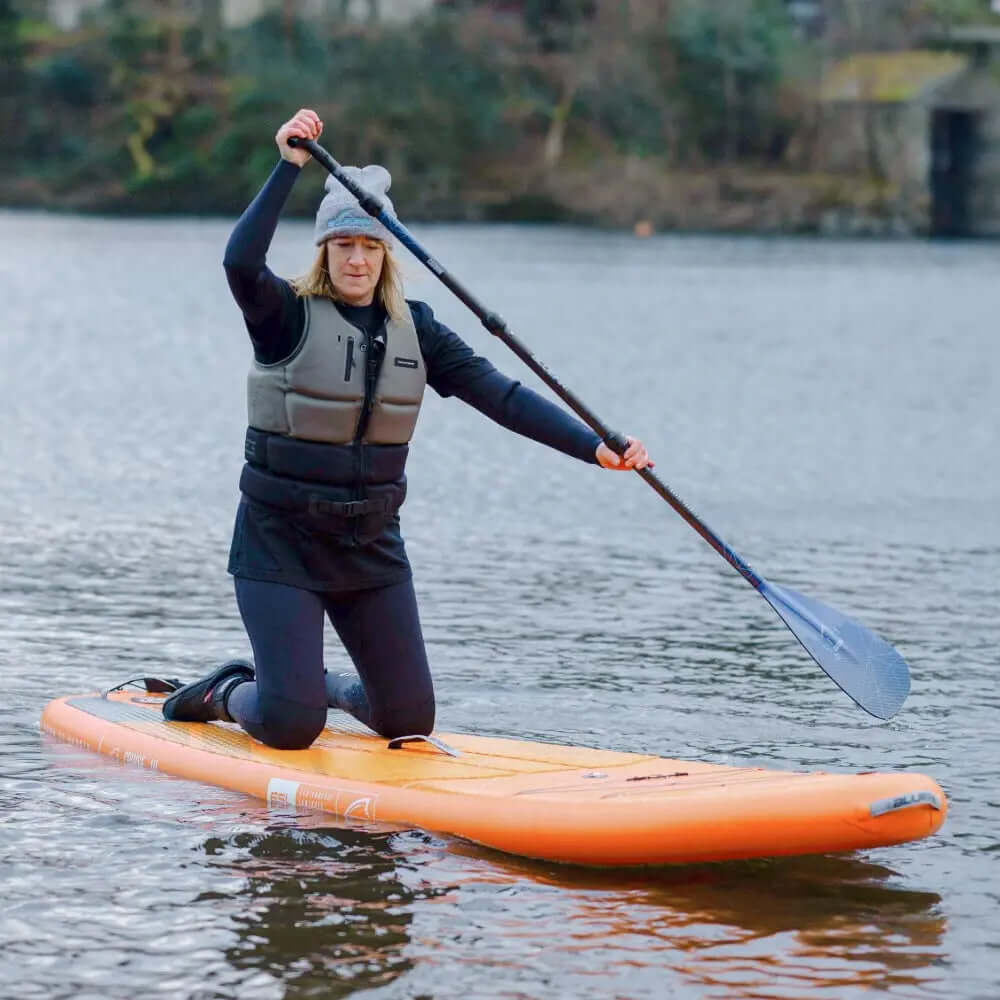 Person kneeling on an orange paddleboard, using a paddle on calm water, wearing a life vest and hat.