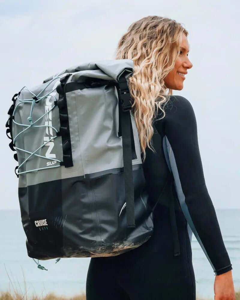 Woman in wetsuit carrying a gray waterproof backpack with blue straps by the beach