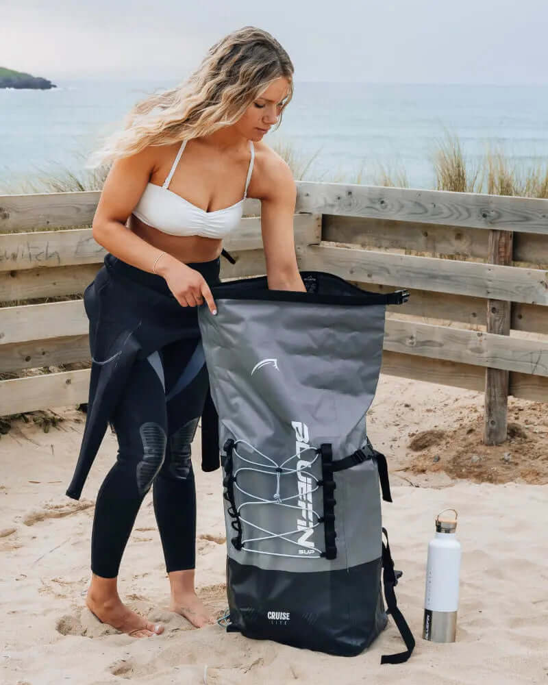 Woman in a bikini and wetsuit opening a gray waterproof backpack on the beach with a water bottle nearby.