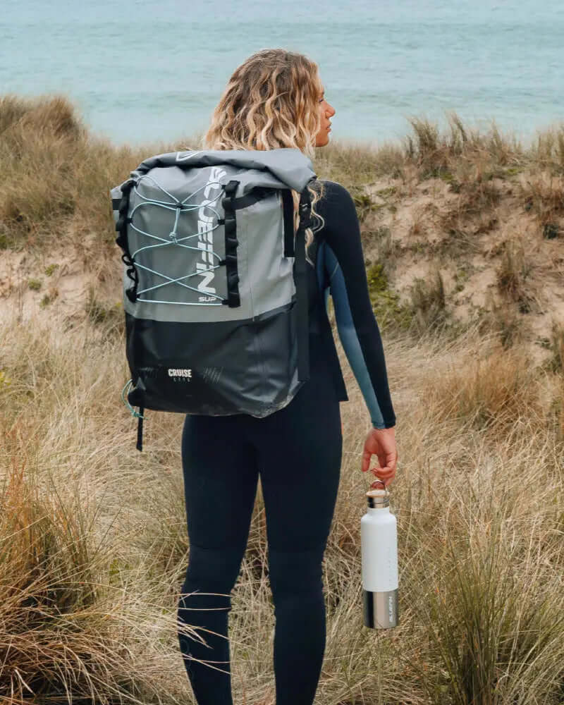 Person in a wetsuit holding a water bottle, wearing a gray backpack by Bluefin SUP near the beach.