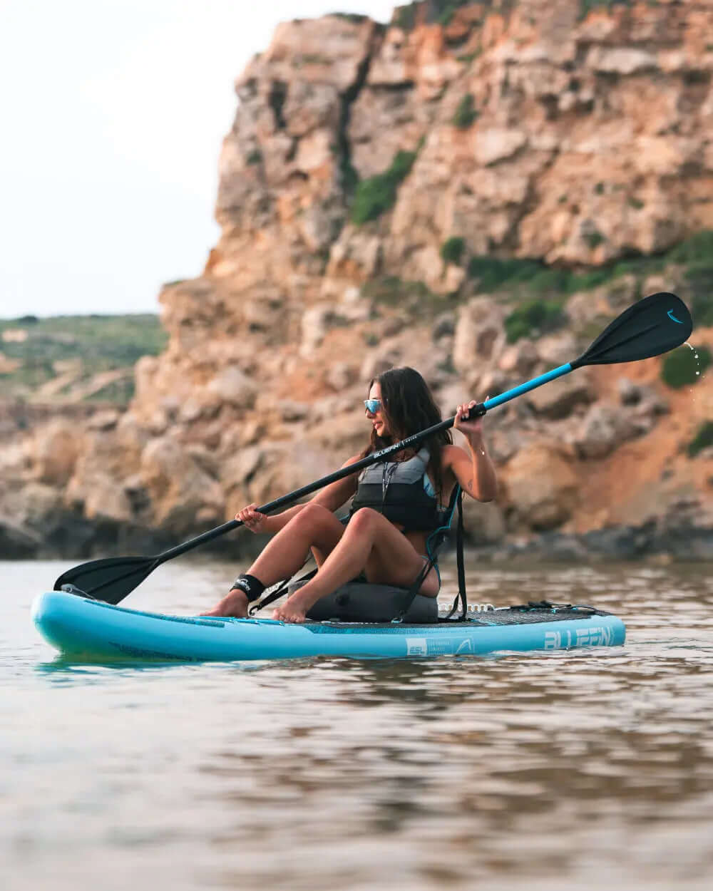 Frau paddelt auf einem blauen aufblasbaren Paddleboard in der Nähe von felsigen Klippen im ruhigen Wasser.