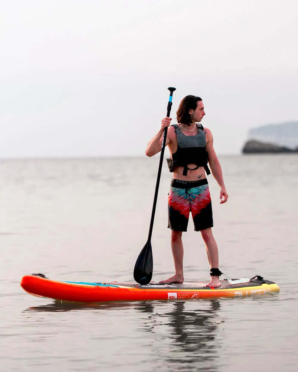 Man on an inflatable paddleboard with a paddle, wearing a life vest and colorful swim shorts