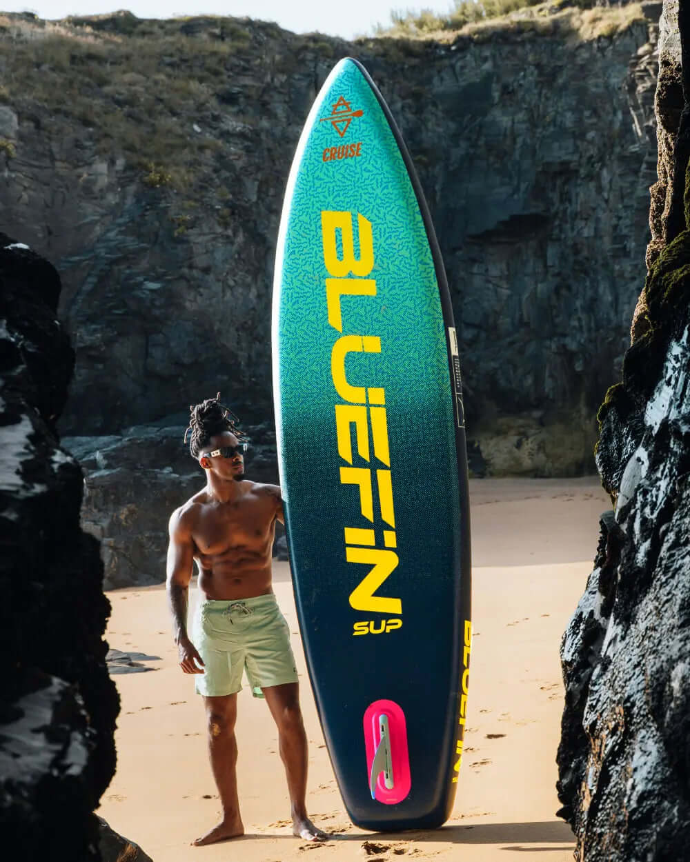 Man standing on beach with Bluefin SUP paddleboard featuring green and yellow design, center fin visible.