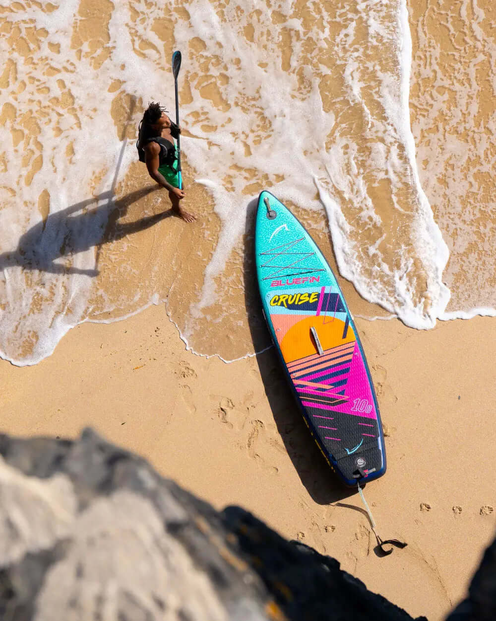 Person standing on sandy beach with paddle, next to colorful Bluefin SUP board in shallow water
