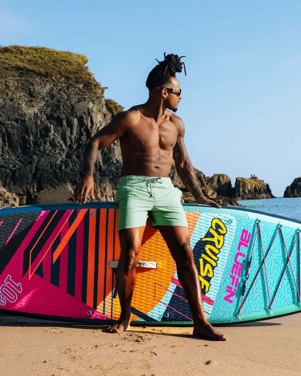 Man in green shorts poses with a colorful Bluefin paddleboard on a sandy beach