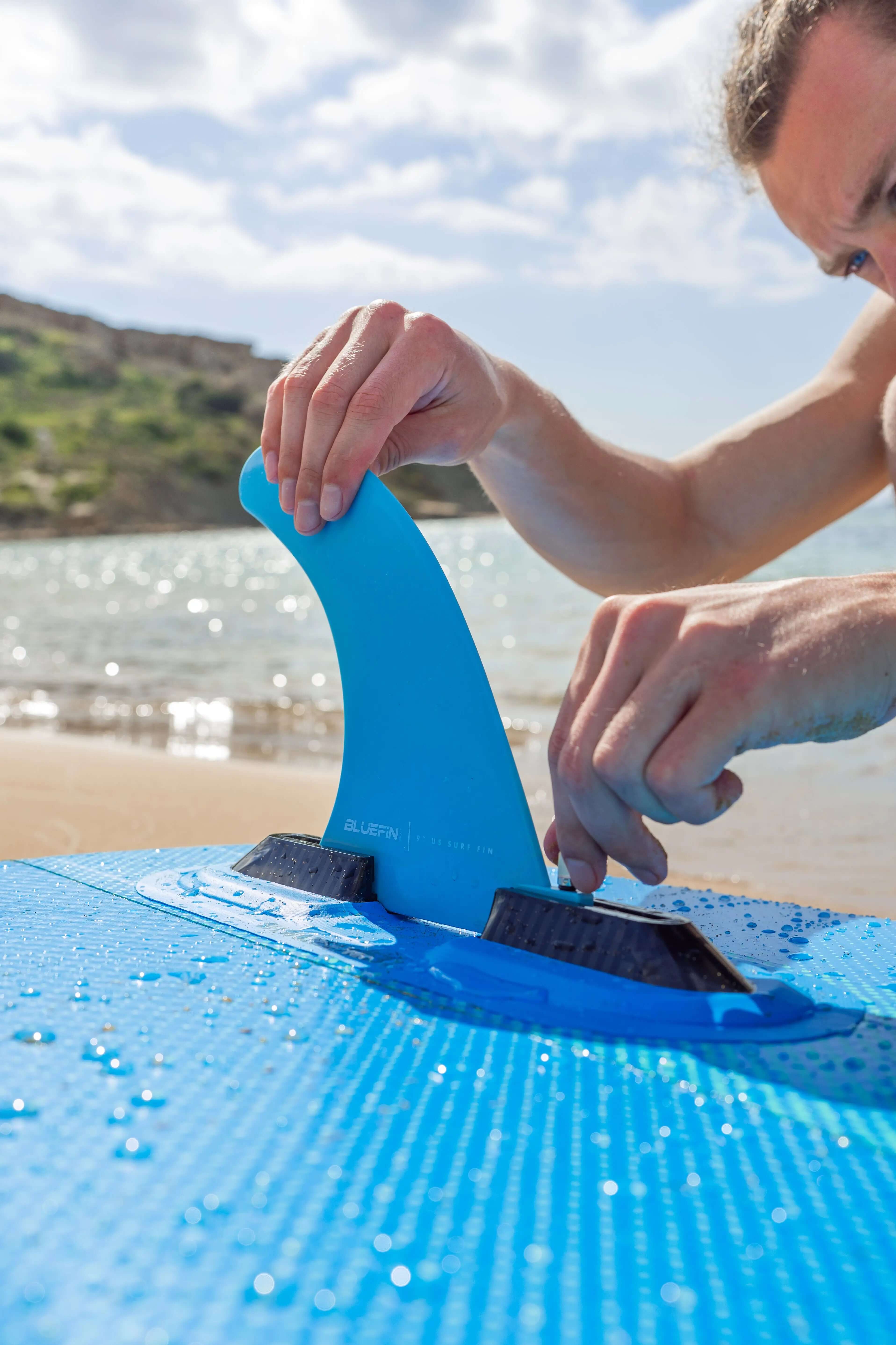 Person attaching a blue fin to a blue paddleboard on a sandy beach with water in the background