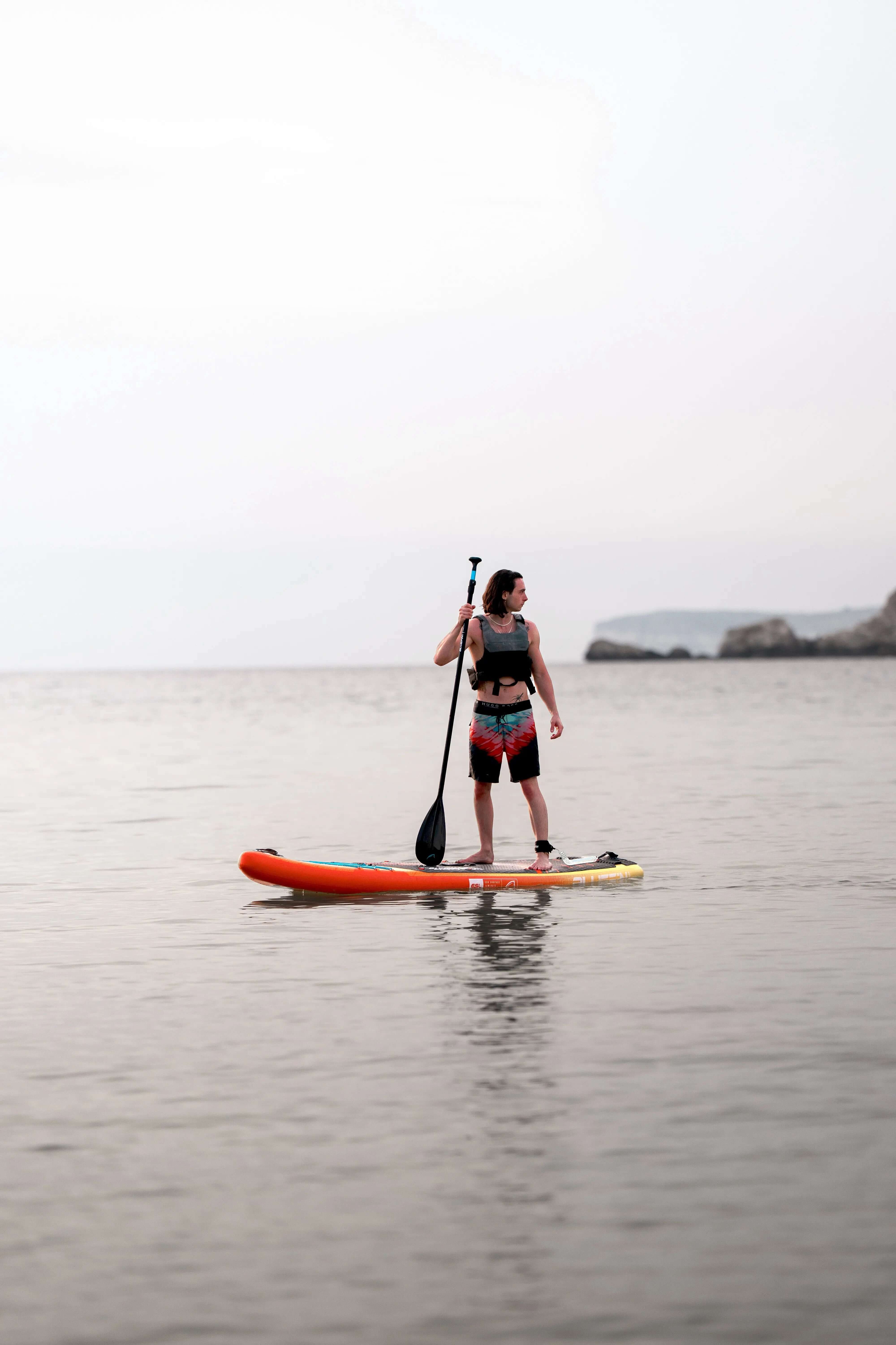 Person on an orange paddleboard in calm water, holding a paddle, wearing swim shorts and a life vest.