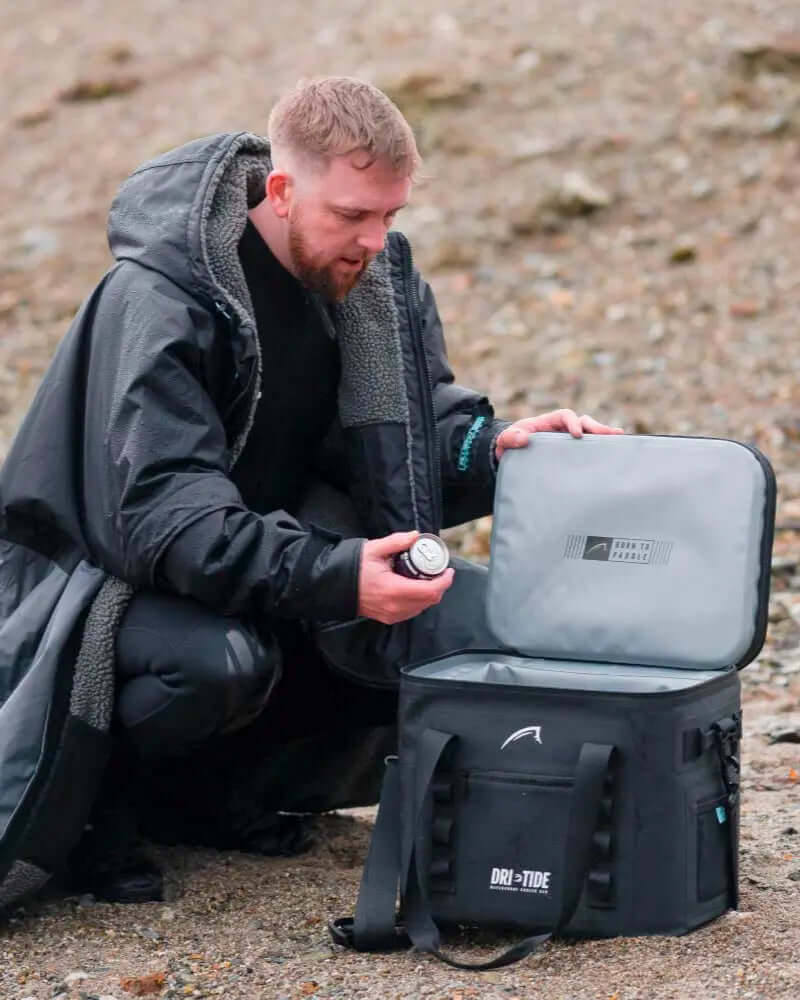 Person in a black coat kneeling by a black cooler bag with an open lid, holding a can.