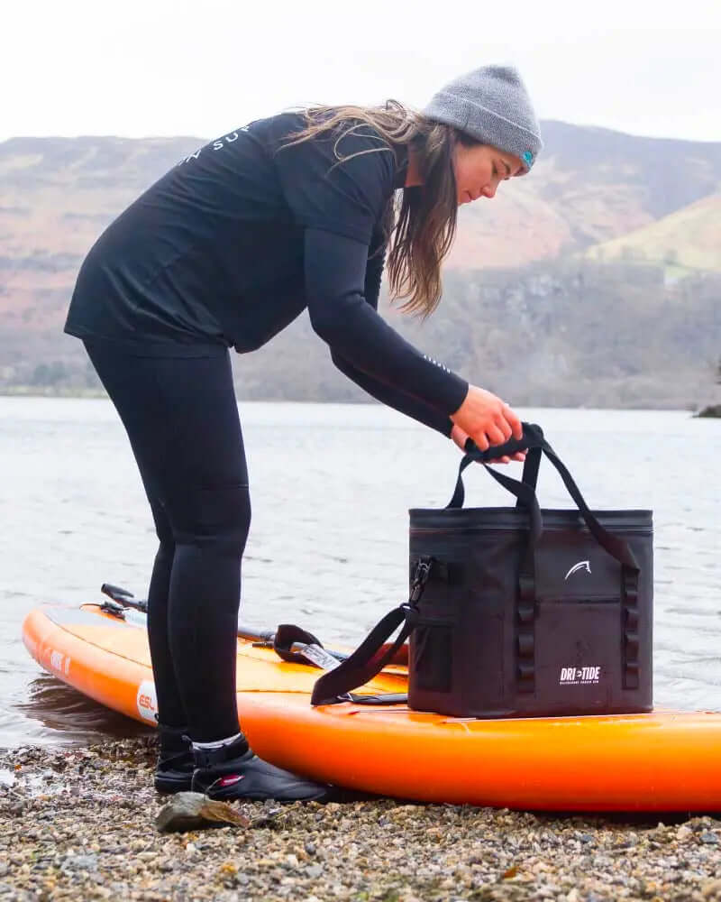 Person in black outfit and beanie lifting a black cooler from an orange paddleboard by the shore