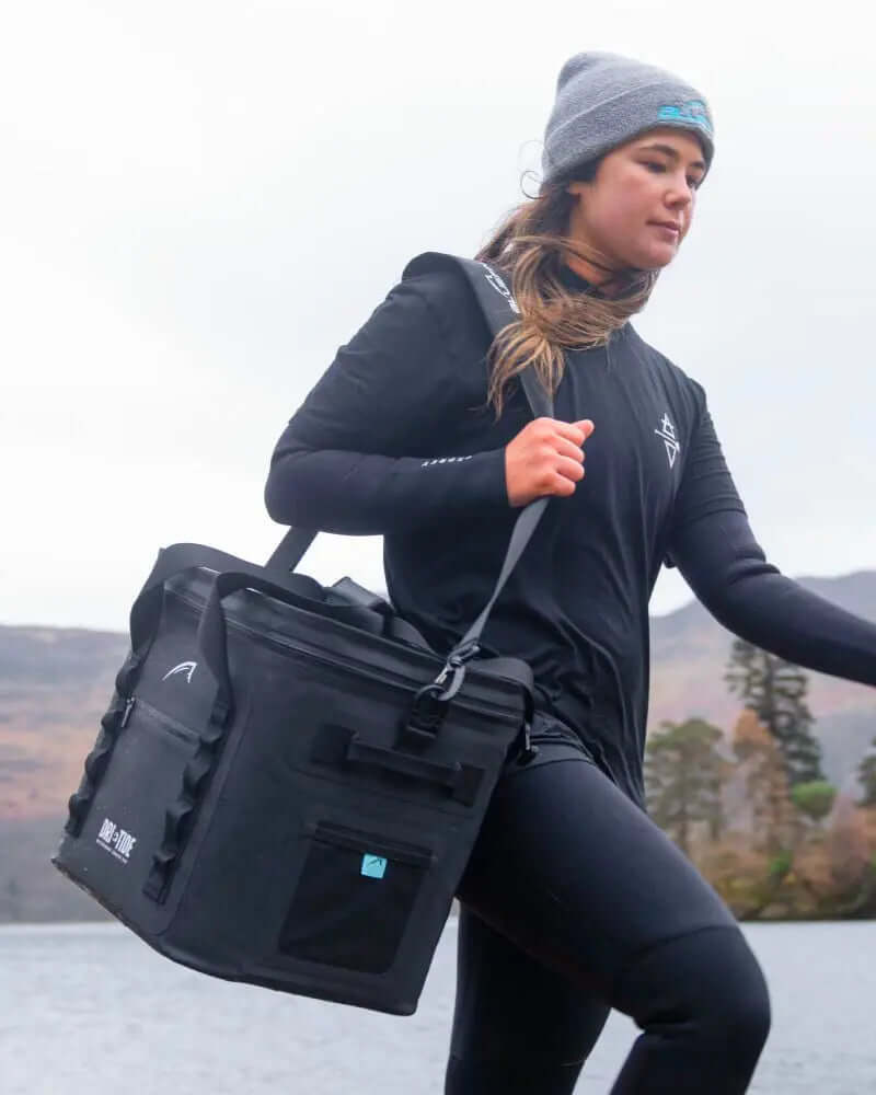 Person carrying a black cooler bag with a shoulder strap near a lake, wearing a long-sleeve shirt and beanie.