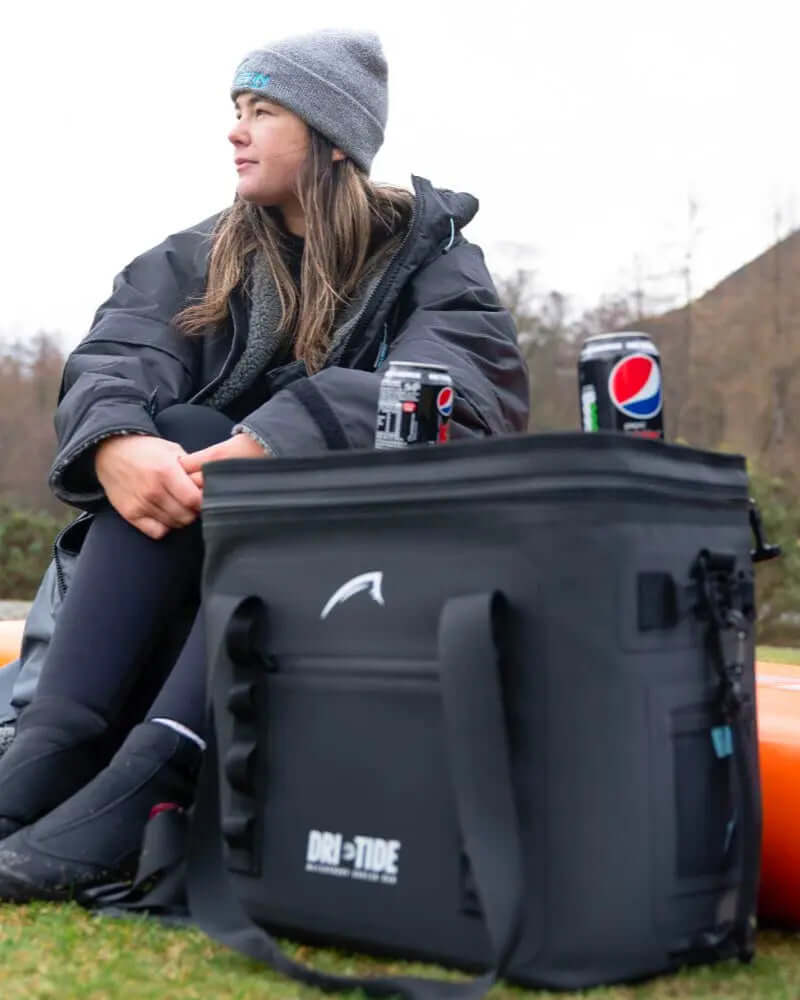Woman in a gray beanie sitting beside a black cooler bag with drinks, wearing a black jacket and wetsuit.