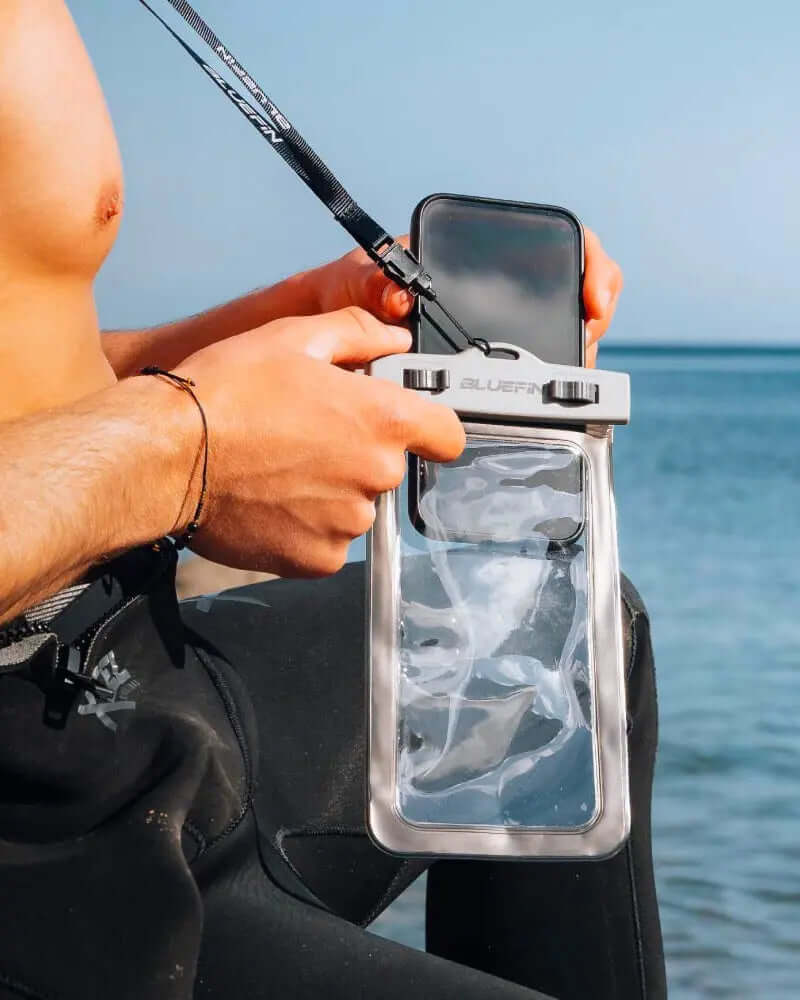 Person holding a waterproof phone case with a lanyard by the water, wearing a wetsuit.