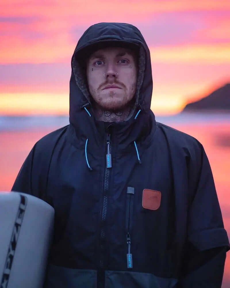 Person in a black hooded jacket standing by the ocean at sunset, with a surfboard beside them.