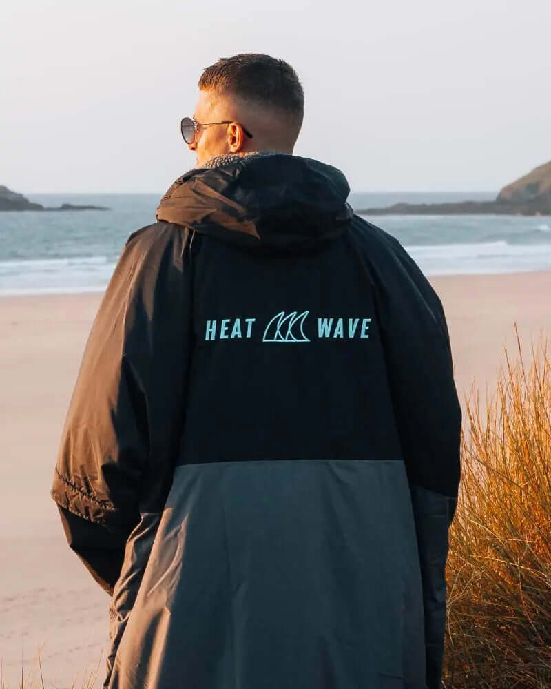 Person wearing a black and gray jacket with 'HEAT WAVE' logo, standing on a beach at sunset.