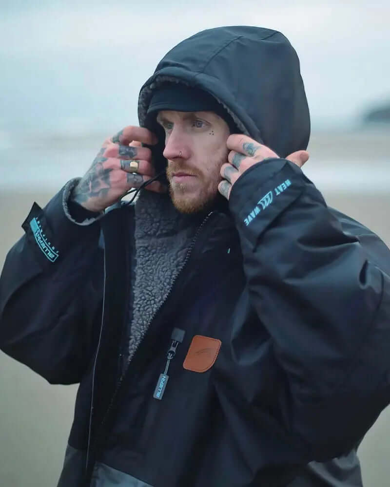 Man in a black waterproof jacket with a hood, adjusting it outdoors near the beach.