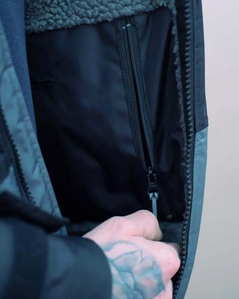 Interior of a jacket showing a black zippered pocket and a hand reaching inside, with a tattoo visible.