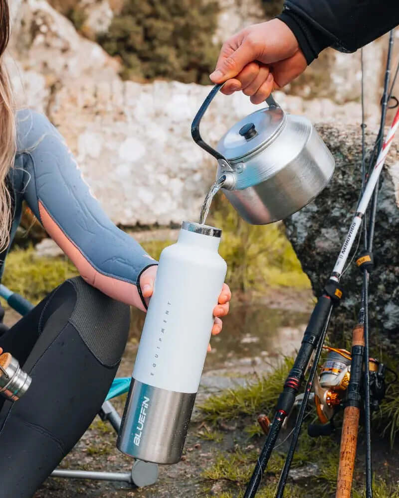 Person in wetsuit pouring water from a kettle into a Bluefin water bottle by fishing rods outdoors.