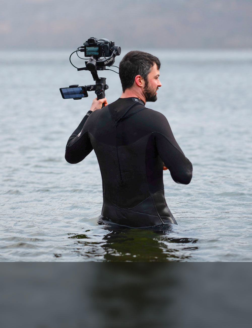 Man in a black wetsuit holding a camera stabilizer while standing in shallow water