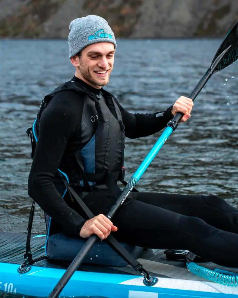 Man in wetsuit paddling on a blue inflatable paddleboard on water, smiling with a paddle in hand.