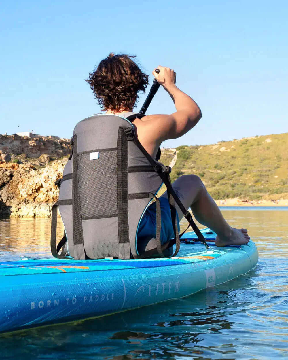 Person paddling on a blue inflatable paddleboard with a gray backrest in calm water.