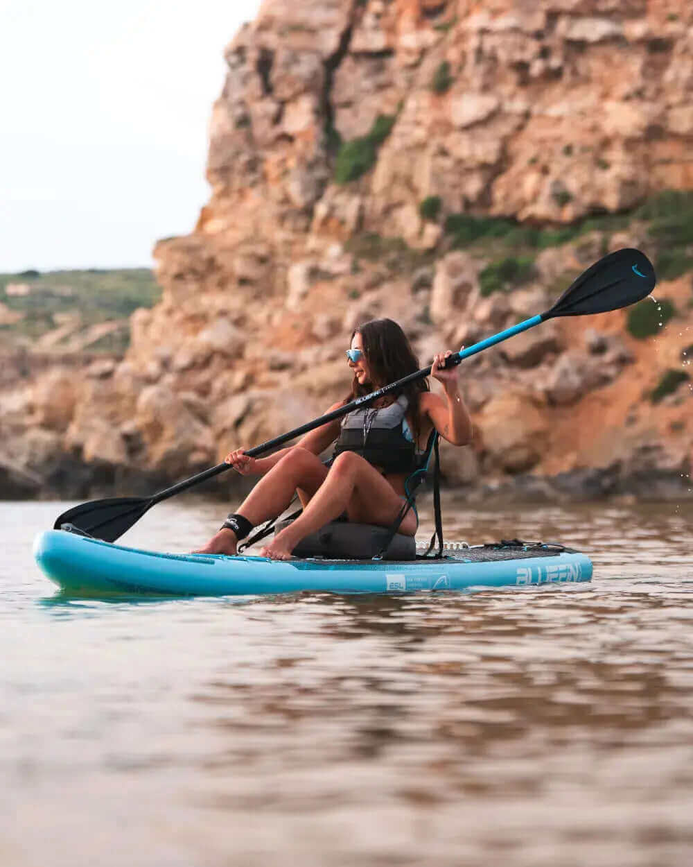 Woman paddling on a blue inflatable paddleboard in calm water near rocky cliffs